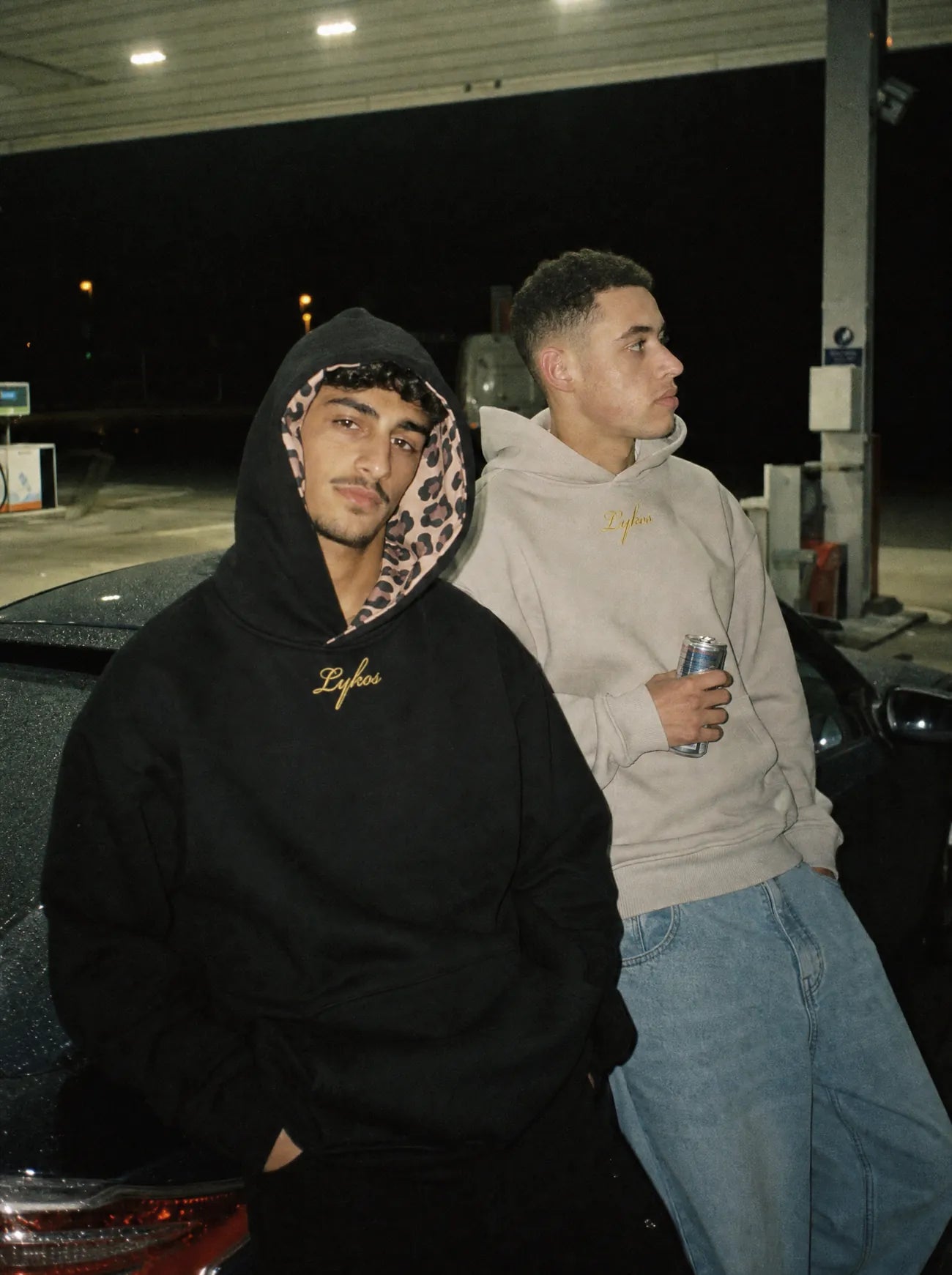 Two friends at a petrol station at night in the Origin Hoodie, black with the hood up and grey