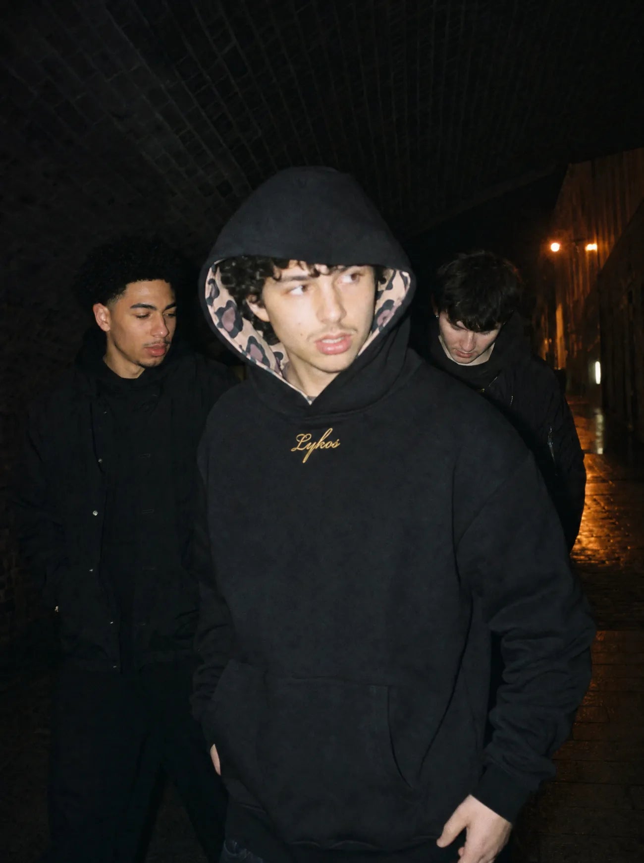 Black Origin Hoodie worn with the hood up under a railway arch at night